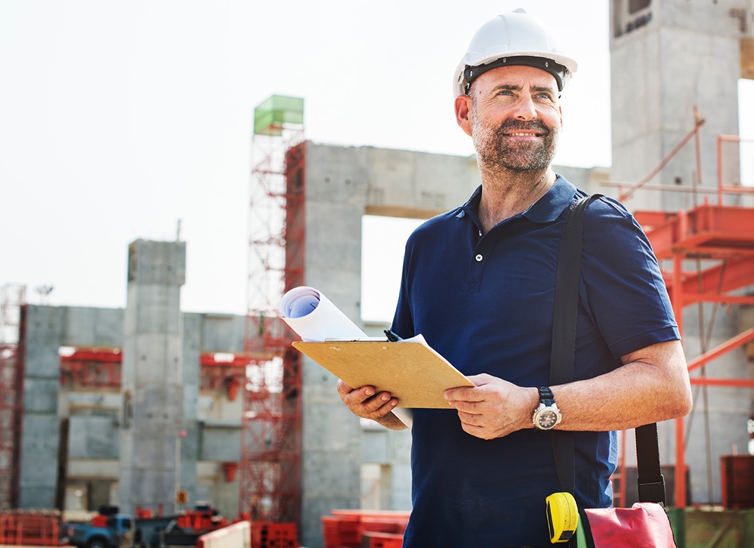 Insurance by Industry - Closeup Portrait of a Smiling Middle Aged Contractor Holding a Notepad and Blueprints While Standing in a New Development Construction Project Build Site